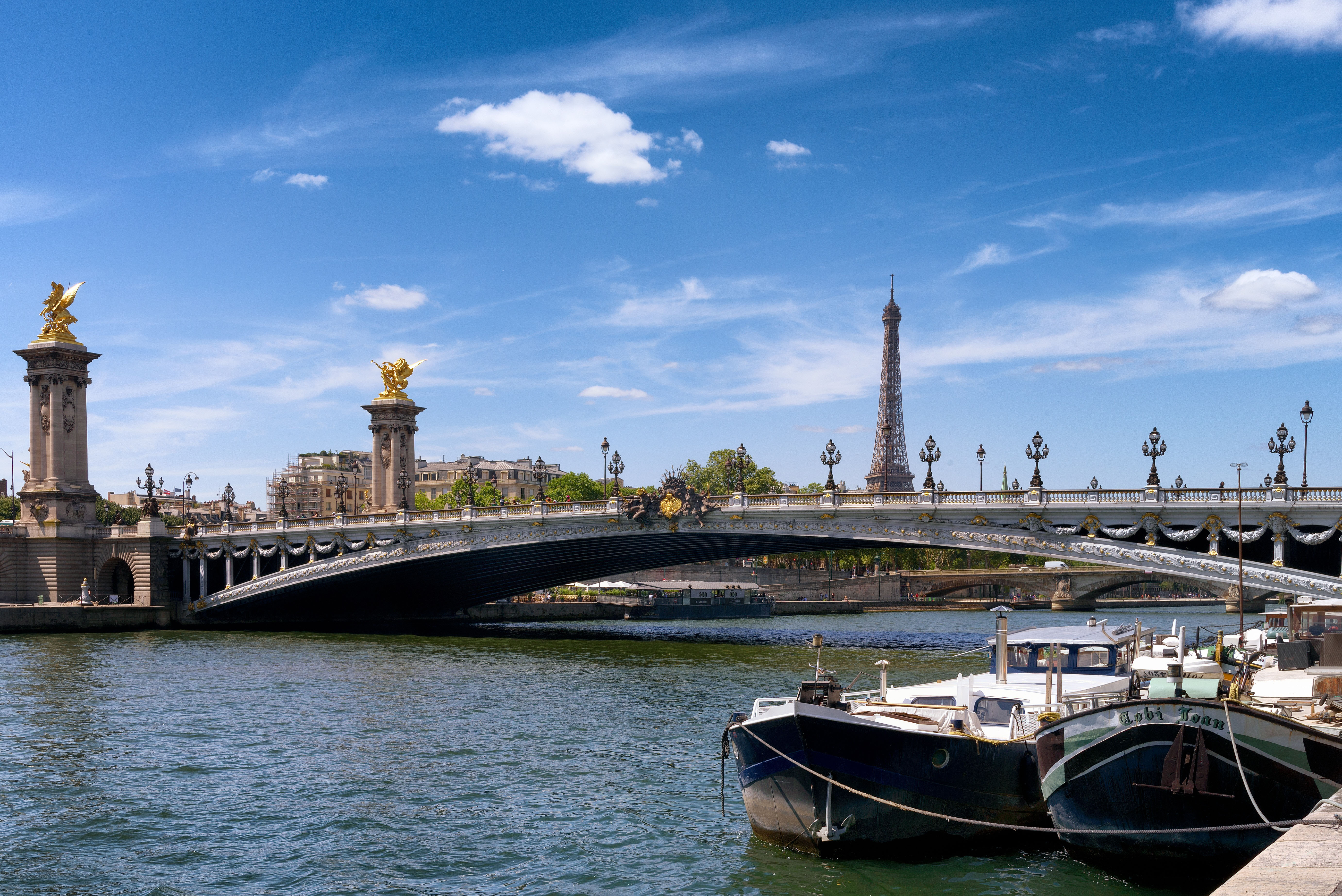 Pont Alexandre III-bron �ver floden Seine i Paris med Eiffeltornet i bakgrunden och b�tar vid kajen under bl� himmel.