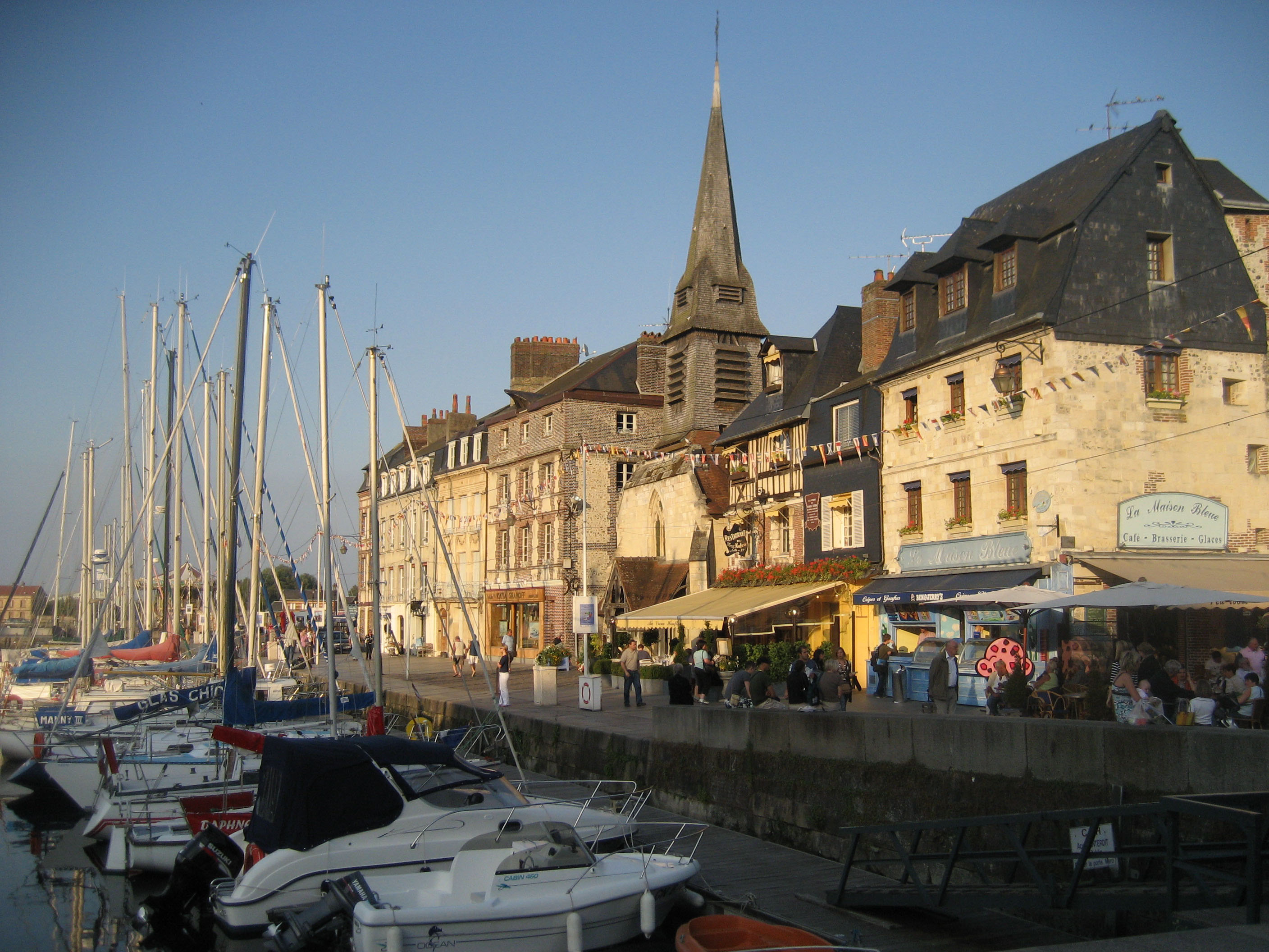 Historiska byggnader och segelb�tar vid hamnen i Honfleur, Normandie, med pittoreska restauranger och kyrkspira under klar himmel.