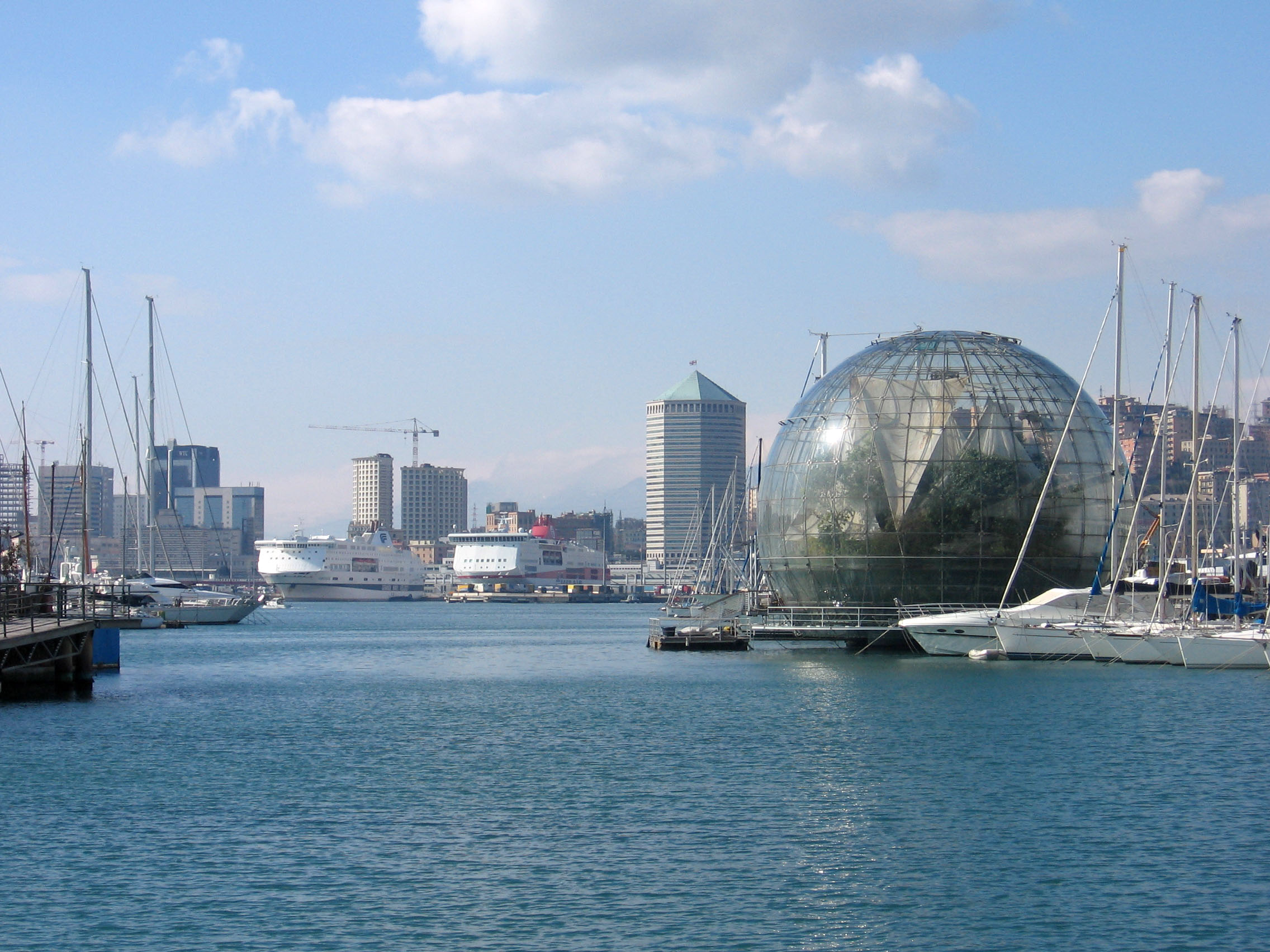 Modern hamn i Genua, Italien med segelbåtar, futuristisk glaskupol och stadens skyline under blå himmel.