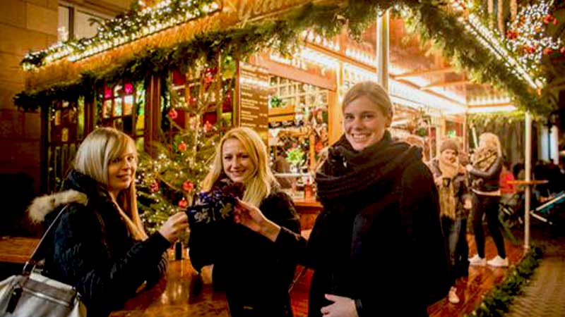 Tjejer p� julmarknadsresa till Tyskland d�r de njuter av v�rmande Gluhwein i ett marknadsst�nd.