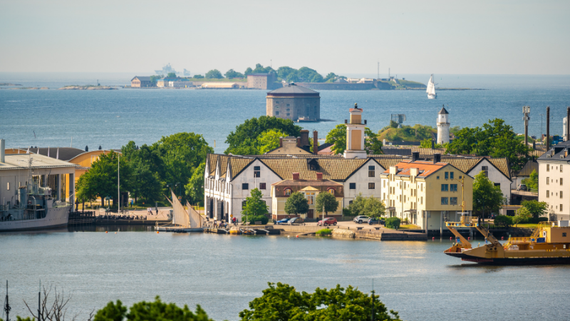 Utsikt �ver Karlskronas historiska varvsomr�de med vackra byggnader vid vattnet, sk�rg�rds�ar i bakgrunden och b�tar som trafikerar hamnen en solig sommardag.
